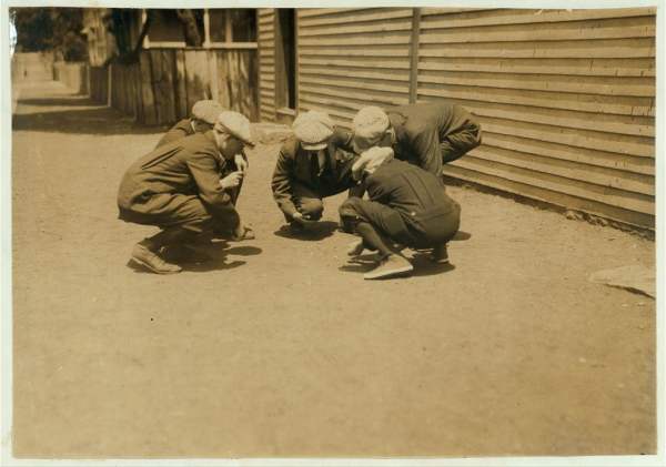 Sepia-toned photograph of boys gambling over cards. They are crouched in a circle on hard packed dirt outside a building.